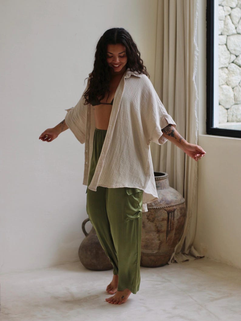 Puede incluir: Una mujer con una camisa beige abotonada de gran tama&ntilde;o, pantalones verde oliva y una parte superior negra. La camisa tiene un aspecto texturizado. Los pantalones son de color liso con un ligero brillo. La mujer sonr&iacute;e y tiene el pelo largo y oscuro.