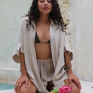 May include: A woman wearing a light beige, loose-fitting shirt and matching shorts. She is kneeling on a white surface with a wooden cutting board in front of her. The cutting board has a variety of fruits on it, including a dragon fruit, a mango, an orange, and a cluster of red rambutan.