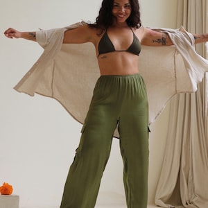 Puede incluir: Una mujer con un top de bikini verde oscuro, pantalones cargo verde oliva y una camisa beige abotonada. Los pantalones tienen bolsillos laterales. La mujer sonr&iacute;e y tiene tatuajes en los brazos.
