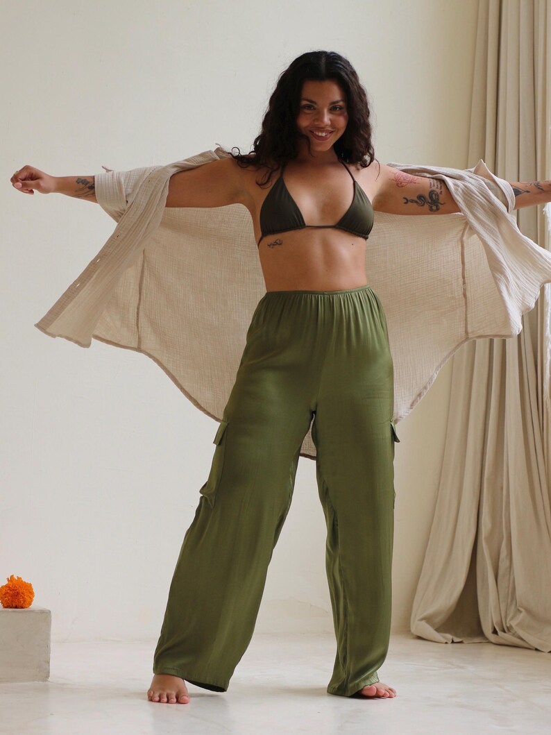 Puede incluir: Una mujer con un top de bikini verde oscuro, pantalones cargo verde oliva y una camisa beige abotonada. Los pantalones tienen bolsillos laterales. La mujer sonr&iacute;e y tiene tatuajes en los brazos.