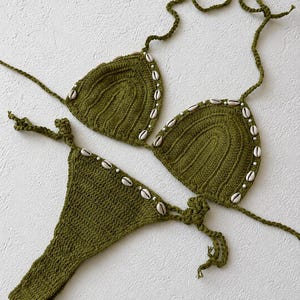 Könnte beinhalten: Ein olivgrünes Häkel-Bikini-Set bestehend aus zwei dreieckigen Cups und einem passenden Unterteil. Die Ränder des Bikinis sind mit kleinen weißen Kaurimuscheln und winzigen weißen Perlen verziert. Das Set enthält dünne gehäkelte Bindebänder.