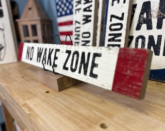 Puede incluir: Un letrero rectangular de madera con un acabado blanco desgastado y extremos pintados de rojo. El texto NO WAKE ZONE está impreso en letras mayúsculas negras, negritas y sin remates en el centro. El letrero se apoya en un soporte de alambre negro.