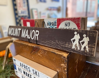 May include: A brown wooden sign with white text that reads "MOUNT MAJOR". Two white stick figures are hiking with hiking poles on the sign. The sign is hanging on a wooden shelf with other signs.