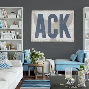 May include: A living room interior with a white couch, blue accent chairs, and a white bookshelf filled with books. A large wooden sign with the text "ACK" in blue letters hangs on the wall. The sign has a whitewashed background and a distressed look.