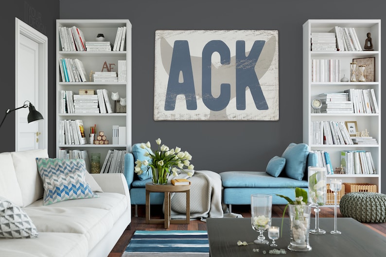 May include: A living room interior with a white couch, blue accent chairs, and a white bookshelf filled with books. A large wooden sign with the text "ACK" in blue letters hangs on the wall. The sign has a whitewashed background and a distressed look.