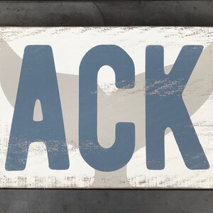 May include: A white wooden sign with a distressed finish featuring the name "ACK" in blue letters. A gray whale tail is behind the letters.