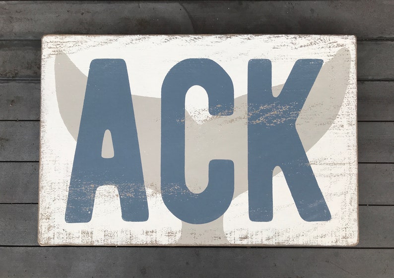 May include: A white wooden sign with a distressed finish featuring the name "ACK" in blue letters. A gray whale tail is behind the letters.