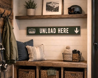 Puede incluir: Un letrero rectangular de madera verde con texto blanco que dice UNLOAD HERE, con un gráfico blanco de un telesilla y una flecha que apunta hacia abajo. El letrero está montado en una pared de listones blancos sobre un banco de madera.