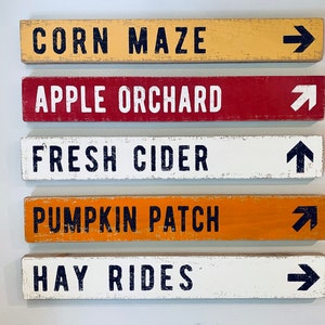 May include: Five wooden signs with white painted text and black arrows pointing to the right. The signs read: "Corn Maze", "Apple Orchard", "Fresh Cider", "Pumpkin Patch", and "Hay Rides".