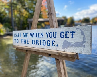 May include: A white wooden sign with blue text that reads "Call me when you get to the bridge." The sign is on a wooden easel and has a gray outline of Cape Cod on the right side.