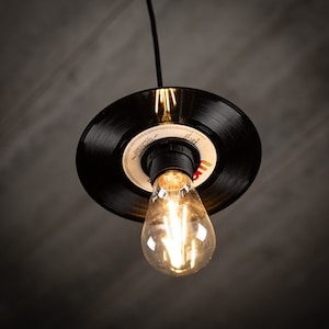 May include: A black vinyl record has been upcycled into a pendant light fixture with a clear light bulb. The light bulb is switched on and illuminating the room.