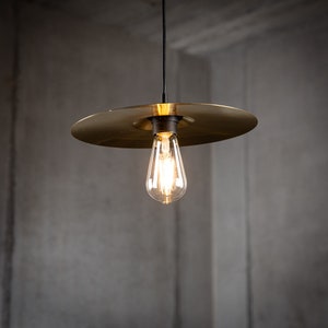Op de afbeelding: Een gouden hanglamp met een zwart snoer en een heldere gloeilamp. De lamp heeft een brede, platte, ronde kap.