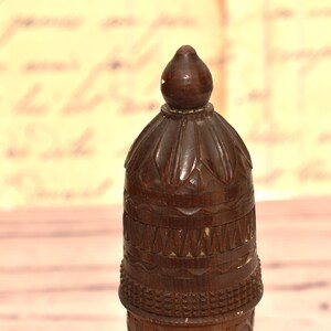 May include: A dark brown wooden bottle stopper with intricate carvings. The top of the stopper is shaped like a dome with a pointed tip.