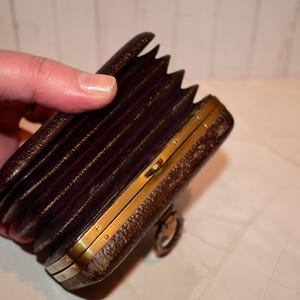 May include: A vintage brown leather camera with a bellows design and brass detailing. The camera is held in a hand, highlighting its accordion-like structure and classic aesthetic. The camera's design suggests it is an antique or collectible item.
