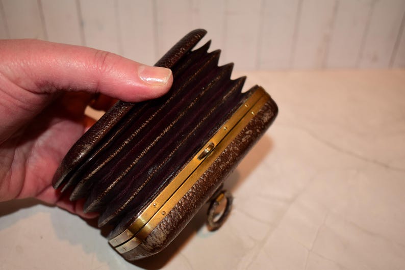 May include: A vintage brown leather camera with a bellows design and brass detailing. The camera is held in a hand, highlighting its accordion-like structure and classic aesthetic. The camera's design suggests it is an antique or collectible item.