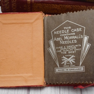Puede incluir: Estuche de agujas vintage abierto con cubierta roja e interior beige. El estuche contiene un papel marrón con el texto "ABEL MORRALL'S NEEDLES" y "MADE IN ENGLAND". El estuche está abierto, revelando las agujas en su interior. Una pila de libros antiguos está en el fondo.