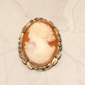 May include: A cameo brooch with a silver tone setting and a cameo of a woman's profile. The setting is decorated with marcasite stones.