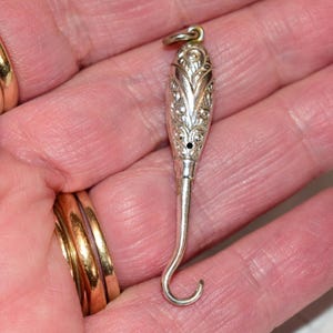 Peut inclure: Pendentif argenté avec un haut orné et allongé et un bas incurvé en forme de crochet. Le haut présente des motifs floraux et de volutes détaillés. Une petite boucle est en haut pour suspendre.