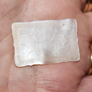 Peut inclure: Pendentif rectangulaire blanc cassé avec un motif floral sculpté. Le pendentif a un bord légèrement irrégulier et un petit trou en haut. Le motif floral est encadré par une bordure décorative. Le pendentif est tenu dans une main.