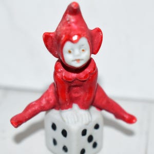 May include: A small figurine of a red-clad elf with a white face and pointed hat, sitting in a split-legged pose atop a white cube-shaped die featuring black pips on its sides. The elf has a smooth, glossy finish and a neutral facial expression.