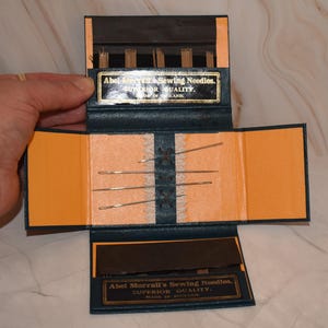 Puede incluir: Un estuche de agujas de coser vintage abierto, con exterior de cuero azul e interior naranja. Contiene agujas y el texto "Abel Morrall's Sewing Needles. SUPERIOR QUALITY. MADE IN ENGLAND."