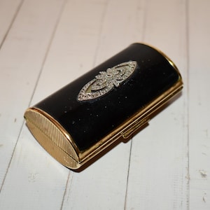 Pode incluir: Um estojo compacto vintage preto e dourado com um adorno decorativo prateado. O estojo compacto tem formato retangular com bordas arredondadas e uma moldura de metal dourado. O adorno é incrustado com pequenas pedras de strass transparentes.