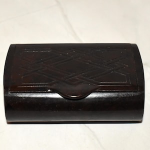 Könnte beinhalten: Eine dunkelbraune, rechteckige Box mit gewölbter Oberseite und Verschluss. Die Box hat ein geometrisches, webartiges Muster auf dem Deckel. Die Box besteht aus einem dunklen, möglicherweise hölzernen Material.