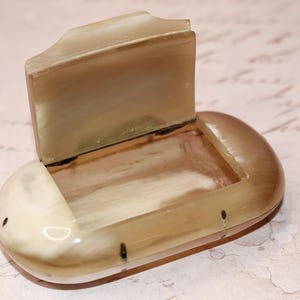 May include: An open, oval trinket box with a hinged lid, made from a light-toned material with a marbled effect. The lid is slightly raised, revealing the interior. The box has a smooth, polished finish.