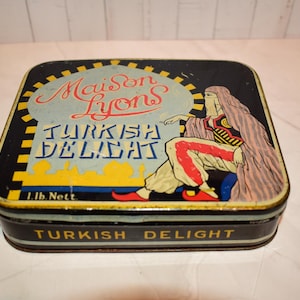 Può includere: Una scatola di metallo rettangolare con sfondo nero e un'illustrazione grafica di una persona in abiti tradizionali. Il testo riporta Maison Lyons Turkish Delight e 1 lb. Nett. I lati sono neri con la scritta Turkish Delight.