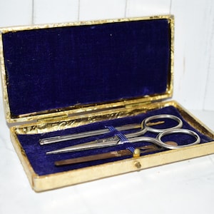 May include: A rectangular gold-coloured case with a hinged lid, featuring a deep blue velvet interior lining. Inside the case, a pair of silver-coloured metal scissors and a thin metal tool are secured in place by a blue elastic band across the centre.