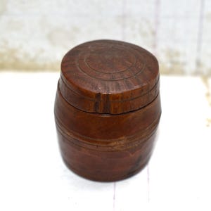 Op de afbeelding: Een klein, tonvormig houten doosje met een deksel. Het doosje is gemaakt van donkerbruin hout en heeft een eenvoudig, rustiek ontwerp.
