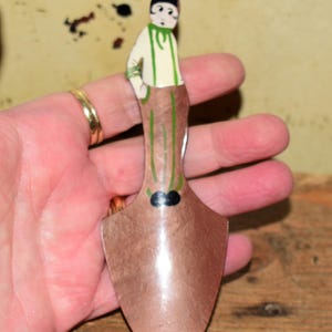 May include: A metal garden trowel featuring a painted figure on the handle. The figure has a black cap, a cream-coloured shirt with green stripes, and brown trousers. The trowel blade is a metallic copper colour with a smooth, reflective finish.