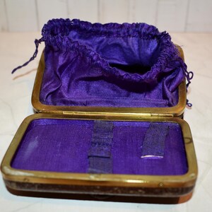 May include: An open antique jewellery box with a brass frame and a deep purple velvet interior. The box contains a matching drawstring pouch. The box is open, revealing the interior and the pouch.