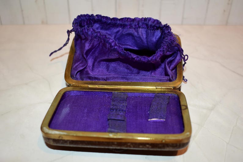 May include: An open antique jewellery box with a brass frame and a deep purple velvet interior. The box contains a matching drawstring pouch. The box is open, revealing the interior and the pouch.
