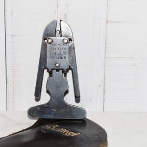 Puede incluir: Una herramienta metálica Buttner con una base en forma de T y una sección superior vertical bifurcada. La placa central presenta el texto en relieve BRITISH BUTTNER PATENT 29243/45. La herramienta descansa sobre una base oscura de cuero con el nombre Buttner.