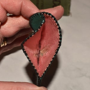 May include: A small, heart-shaped pouch with a green outer layer and a pink inner layer. The pouch is folded in half and the edges are stitched with black thread.