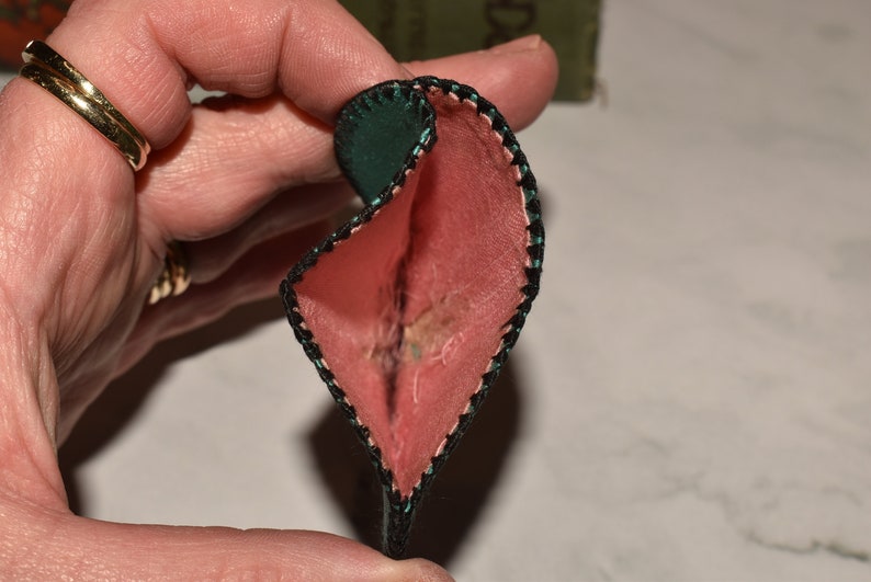 May include: A small, heart-shaped pouch with a green outer layer and a pink inner layer. The pouch is folded in half and the edges are stitched with black thread.