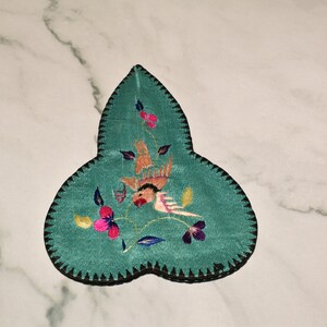 May include: A teal blue fabric leaf-shaped pincushion with black stitching around the edges. The pincushion is embroidered with a pink and orange bird, green leaves, and pink, yellow, and purple flowers.