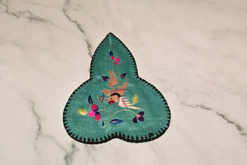 May include: A teal blue fabric leaf-shaped pincushion with black stitching around the edges. The pincushion is embroidered with a pink and orange bird, green leaves, and pink, yellow, and purple flowers.