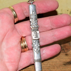 May include: A long, cylindrical silver-toned metal case featuring an embossed design of a central shield topped with a crown, flanked by vertical leafy vine motifs. The item includes a small circular loop at the top for attachment or hanging.