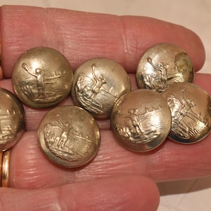 Könnte beinhalten: Sieben silberfarbene Knöpfe mit einem erhabenen Design einer Person, die einen Bogen und Pfeil hält. Die Knöpfe sind in einem Halbkreis auf einer Hand angeordnet.