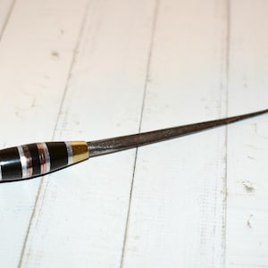 Può includere: Un uncinetto antico in metallo con un manico decorativo. Il manico presenta fasce alternate di nero, argento e rame, con accenti dorati. L'uncinetto stesso è curvo e realizzato in metallo scuro.