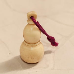 Könnte beinhalten: Ein kleines, kürbisförmiges Dekorationsobjekt in hellbeiger Farbe mit einer dunkelvioletten Kordel. Das Objekt hat eine glatte, polierte Oberfläche und eine geknotete Kordel zum Aufhängen oder Ausstellen.