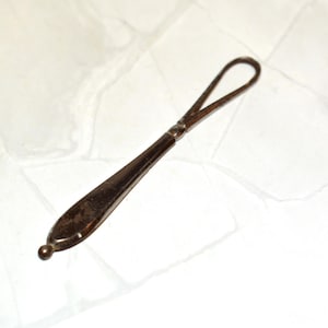 May include: A vintage silver butter spreader with a loop handle. The spreader has a teardrop-shaped blade and a small, round ball at the end. The handle is a darker colour than the blade.