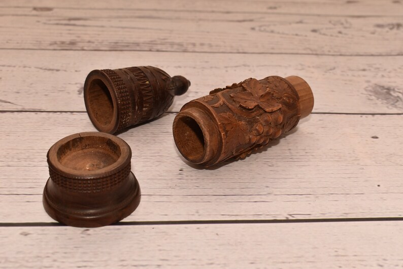 May include: Three wooden containers with intricate carvings. One container is cylindrical with a grapevine design, another is a small round container, and the third is a tall, cylindrical container with a geometric pattern.