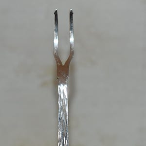 Könnte beinhalten: Eine silberne Serviergabel mit zwei Zinken. Der Griff hat ein gedrehtes Design. Die Gabel besteht aus einem glänzenden, reflektierenden Metall, wahrscheinlich Edelstahl. Der Hintergrund ist eine neutrale, helle Farbe.
