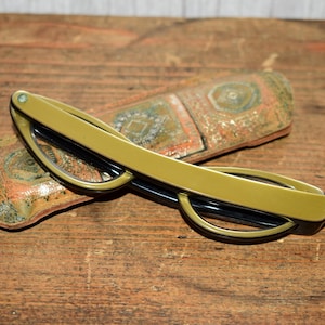 May include: Olive green eyeglasses with black frames rest on a patterned case. The glasses have a rectangular shape and a subtle, vintage aesthetic. The case features a mix of orange, green, and brown tones.