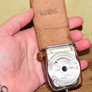 May include: A vintage Sekonic Automat-A light meter housed in a brown leather case. The device features a circular silver dial with numerical scales, a central blue button, and a clear plastic pointer arm. The leather case has a flap with the embossed brand name.