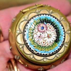 May include: A round, antique gold-toned locket with a mosaic floral design. The mosaic features a central cluster of flowers in pink, white, and blue, surrounded by a turquoise border. The locket is held open, revealing the intricate details.