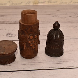 May include: Three wooden containers with intricate carvings. The largest container is cylindrical with a lid and features a grapevine design. The smallest container is bell-shaped with a pointed top. The middle container is a round lid for the largest container.